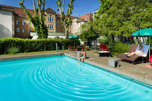 Mann sitzt am Beckenrand des Außenschwimmbades und Frau liegt auf einer Liege im Hintergrund das Bildungshaus St. Bernhard