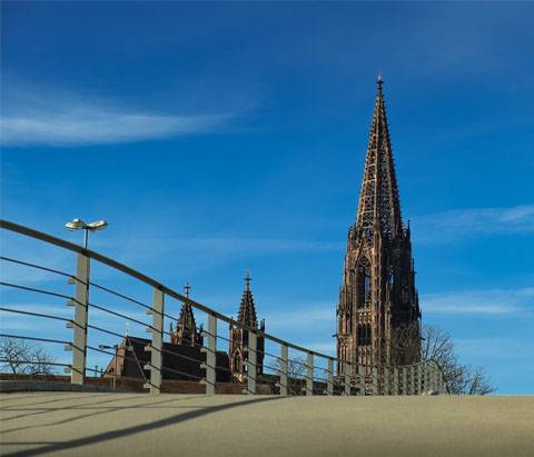 Im Vordergrund ist eine Brücke mit Geländer und im Hintergrund der Turm des Freiburger Münsters vor blauem Himmel und einzelnen Wolken.